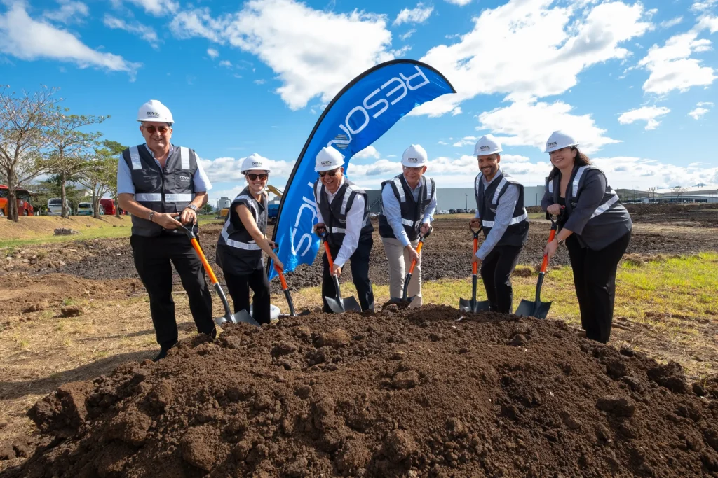 Seis personas con cascos y chalecos de seguridad posan con palas durante una ceremonia de colocación de la primera piedra en un terreno de construcción.