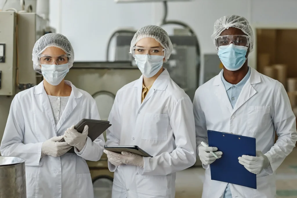 Tres profesionales de laboratorio con batas, guantes, mascarillas, gafas y cofias sostienen tabletas y un portapapeles en una instalación industrial.